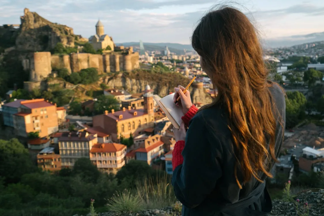 Woman journaling on a top of mounting with beautiful view over the city. Journaling helps reduce stress and anxiety. Journaling helps you track your goals and progress.